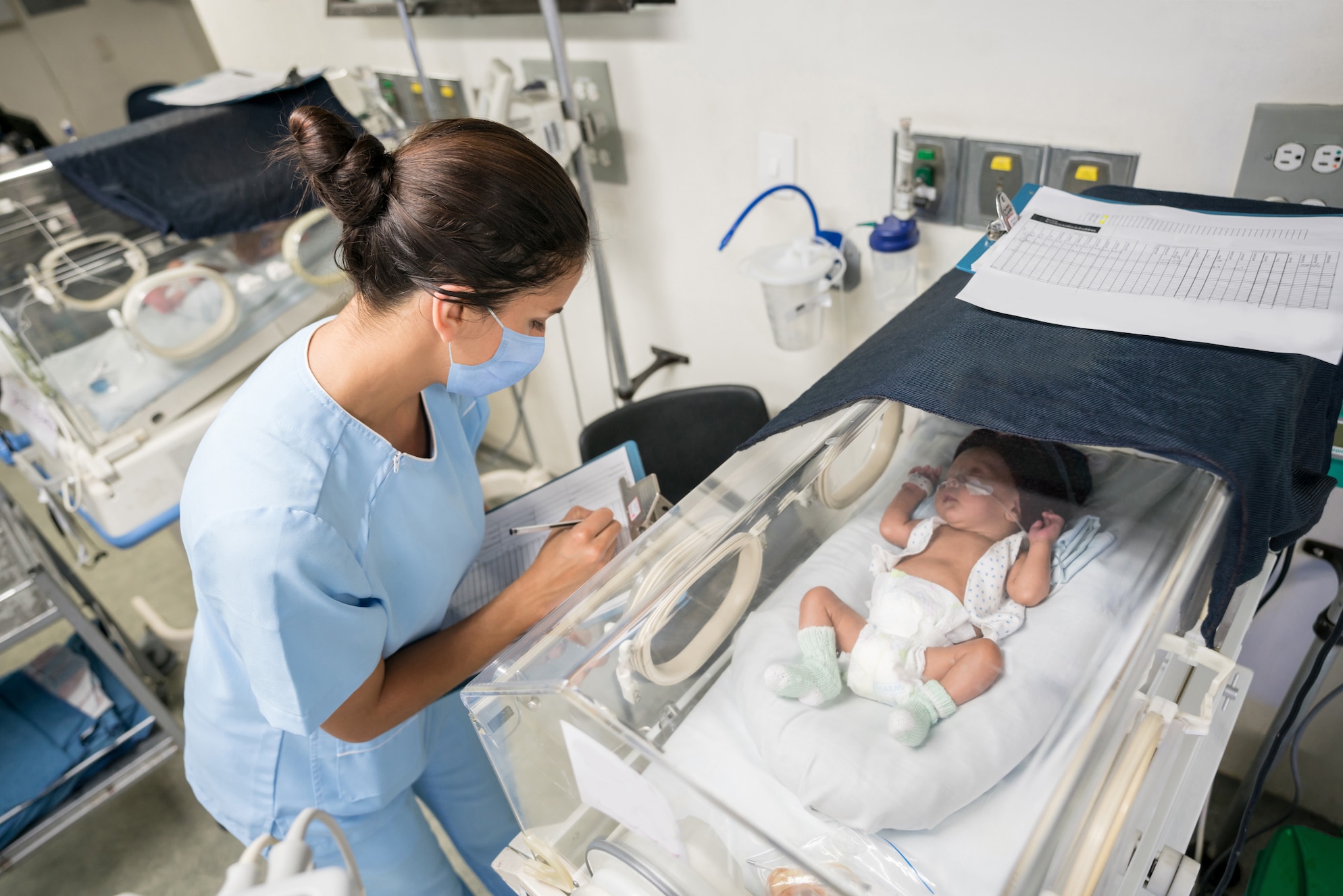 Latin American nurse checking on a premature newborn in an incubator while wearing a facemask during the COVID-19 pandemic